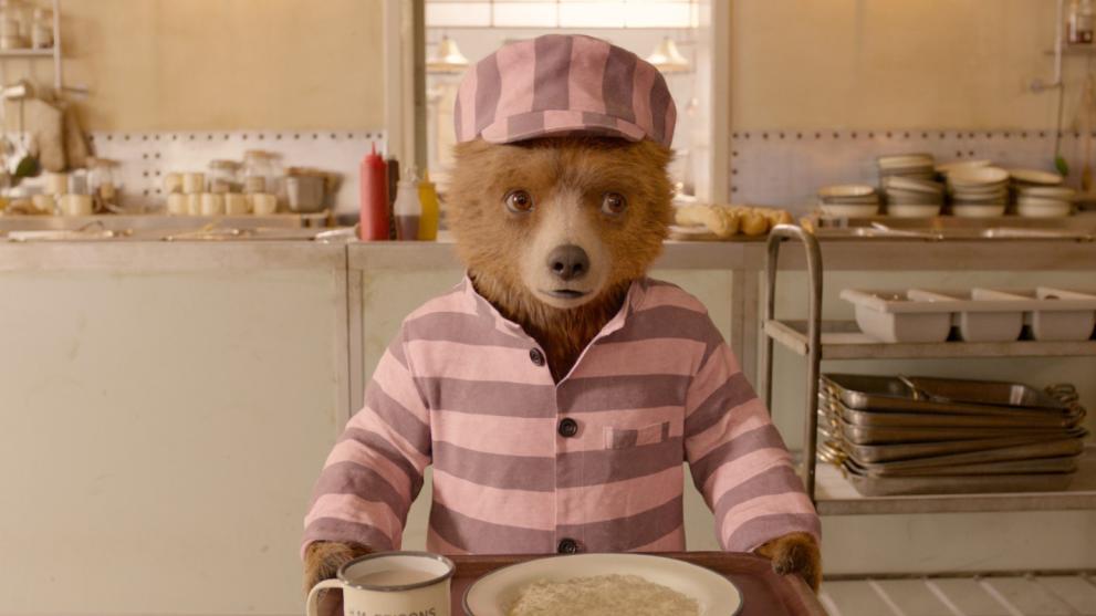 Paddington Bear wearing a pink striped prison uniform, holding a dinner tray in a kitchen.